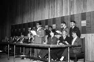 JK discursando durante a sessão de instalação do Congresso enquanto Israel Pinheiro e Oscar Niemeyer acendem seus cigarros; tudo registrado em áudio, em notas taquigráficas e na fotografia de Mario Fontenelli. Ao fundo, a composição dos cartazes do evento<br />Foto Mario Fontenelli 